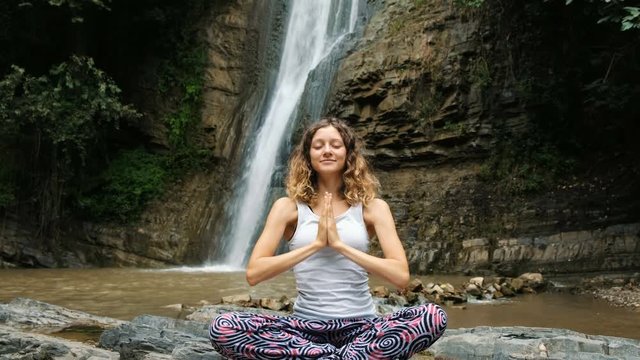 Woman sits in a lotus position and folds her hands in a namaste pose against of a large waterfall.