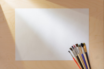 Obraz premium Artist Paint Brushes with Blank White Paper Lying on Wooden Table Illuminated by the Sun. Top View. Drawing Tools in Art Studio.