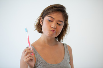 Doubtful pretty woman looking at toothbrush. Young Asian woman holding tooth brush. Teeth hygiene concept. Isolated front view on white background.