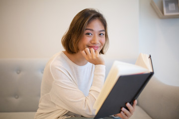 Smiling young woman reading book on sofa at home. Pretty Asian lady looking at camera, sitting and...