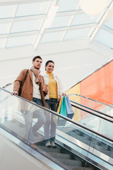 attractive girl with shopping bags and handsome man with disposable cup going down on escalator in mall