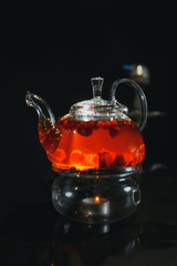 Glass teapot filled with flowers and berries, green tea. Appetizing beautiful drink in orange tones on a black background. Close up.