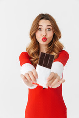 Portrait of excited woman 20s wearing Santa Claus red costume smiling and eating chocolate bar, isolated over white background