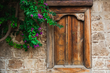 Beautiful wooden closed window in stone wall of old house and green tree with purple flowers growing near it. Horizontal color photography.