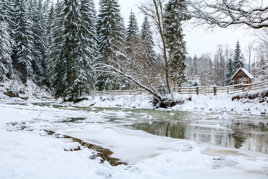 frozen mountain river in spruce snowy forest, fence and arbor on the bank