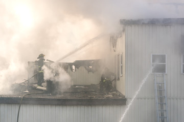 Two unidentifiable firemen fighting a fire from the roof of a building. One sprays water, another holds a hose in a window. Lots of thick smoke. Both wear breathing apparatus.