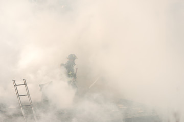 An unidentifiable  fireman on the roof of a burning building. He is surrounded by thick smoke. Smoke is mixed with water spray. Fireman holds an axe. Ladder to the left. Room for text.