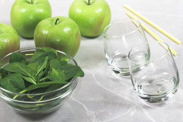 Bowl with fresh spinach leaves, apples, empty glasses and straws. Concept of helathy preparation of smoothie or shake.