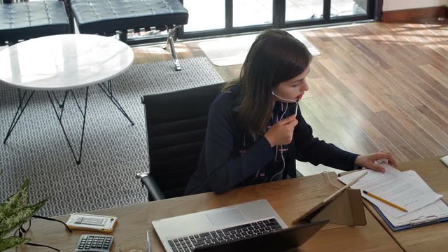 High angle view ofbusiness lady sitting at working table, using headphones for video call, comparing some information in tablet and in papers,  putting everything away and starting typing on laptop
