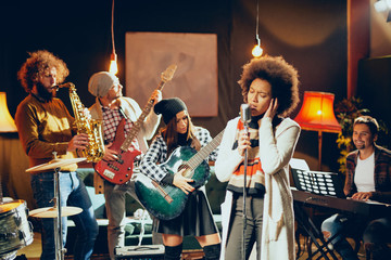 Fototapeta premium Mixed race woman singing. In background band playing instruments. Home studio interior.