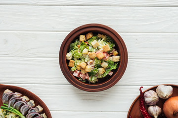 top view of delicious traditional caesar salad with croutons and dishes around on white wooden background