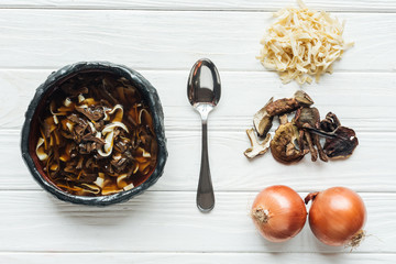 top view of traditional mushroom soup with spoon and ingredients on white wooden background