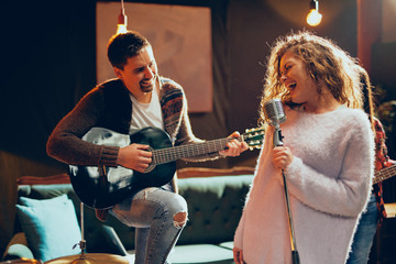 Band practice for the show. Woman with curly hair holding microphone and singing while man in background playing acoustic guitar. Home studio interior.