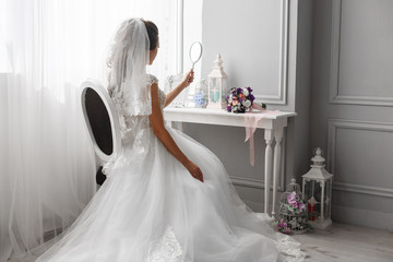 the bride is sitting on the armchair near the window
morning of the bride in a stylish hotel room
beautiful bride looks in the mirror