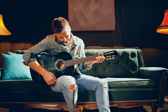 Young bearded focused Caucasian man sitting on the sofa and playing acoustic guitar. Home studio interior.