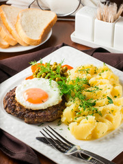 Beefsteak with fried egg and mashed potatoes on white plate close-up on dark brown wooden table in restaurant. Vertical