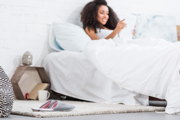 selective focus of slippers on floor and african american woman using digital tablet in bed behind at home
