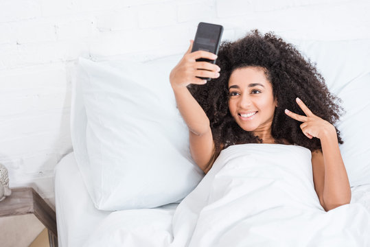 curly african american woman doing victory sign and taking selfie on smartphone