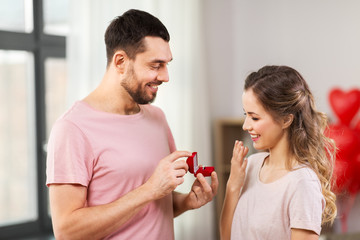 love, couple, proposal and people concept - happy man giving diamond engagement ring in little red box to woman at home