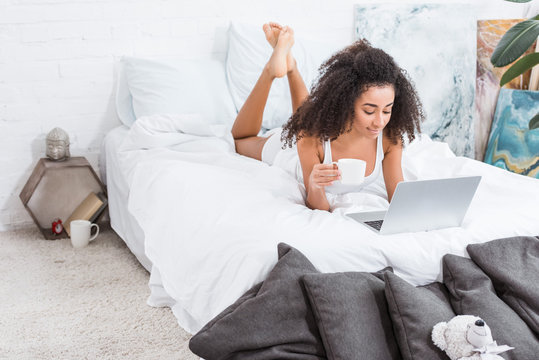 african american curly girl holding coffee cup and using laptop in bed during morning time at home