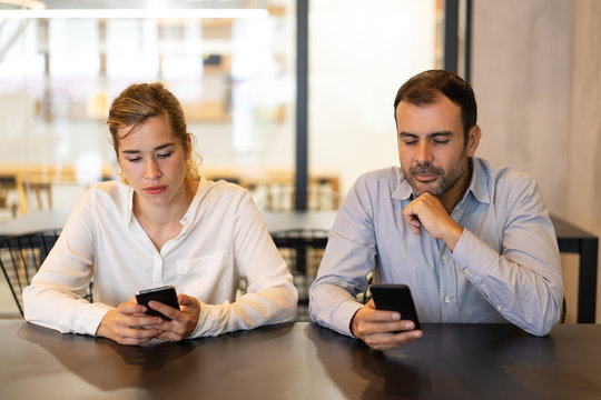 Portrait of male and female colleagues using phones at cafe. Young Caucasian businesswoman and mid adult bearded businessman sitting together at table and networking on smartphones. Addiction concept