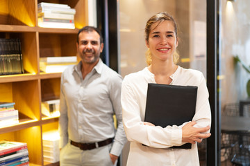 Portrait of successful businesswoman standing with folder in her office, mid adult male colleague...