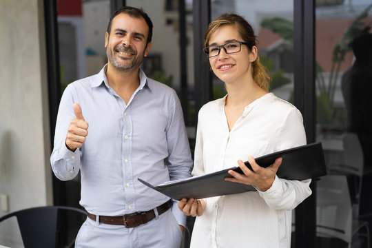 Portrait of successful team of young woman and mid adult man. Businessman smiling and showing thumbs up, woman wearing glasses holding folder. Success concept