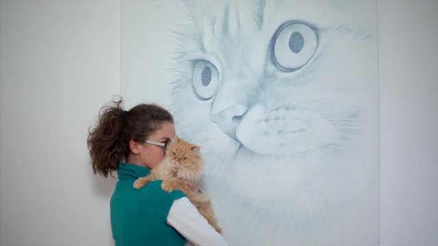 the girl holds a red cat in her arms, the cat bites the hostess by the nose. the girl stands on the background of a picture of a cat.