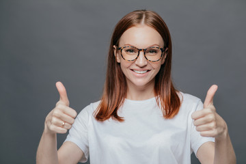 Glad positive woman with tender smile on face, has brown hair, raises two thumbs, demonstrates her approval, isolated over grey background, wears spectacles, wears casual white t shirt. Body language