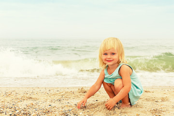 Fototapeta na wymiar A little girl with bright hair plays with the sand on the sea beach.