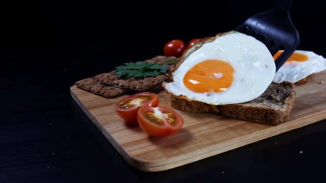 Healthy breakfast. A man is putting scrambled eggs on a fresh grain bread. There are ripe cherry tomatoes, bread crips with parsley on the cutting board. Camera moves from left to right