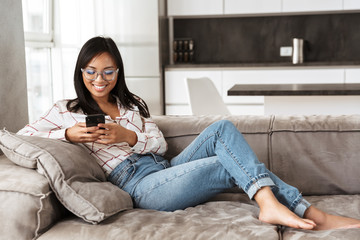 Photo of gorgeous asian woman 20s holding mobile phone, while lying at sofa in cozy flat