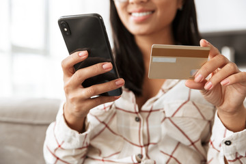 Photo of smiling asian woman 20s holding credit card and mobile phone, while sitting at sofa in apartment