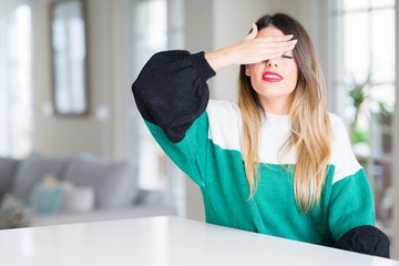 Young beautiful woman wearing winter sweater at home smiling and laughing with hand on face covering eyes for surprise. Blind concept.