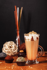 Beautifully designed latte macchiato coffee with whipped cream and sprinkled with crushed halva, in high transparent glass, on a wooden table, on a dark background with cakes