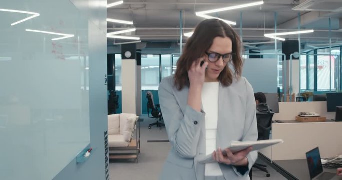 TRACKING Mid 30s Caucasian woman executive walking through busy office, talking on the phone. 4K UHD - Powered by Adobe