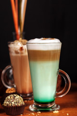 Coffee latte macchiato with mint syrup and whipped cream, in high transparent glasses, on a wooden table, on a dark background with cakes