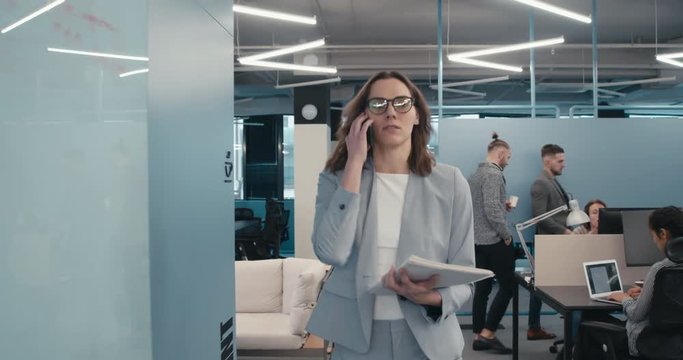 TRACKING Mid 30s Caucasian woman executive walking through busy office, talking on the phone. 4K UHD
