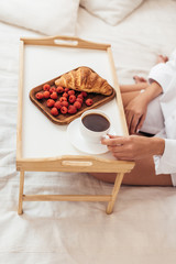 cropped image of young woman holding coffee cup while sitting on bed with breakfast on tray