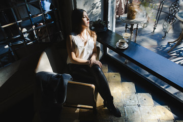 Urban female portrait. Attractive young brunette woman dressed in casual style sits on the chair before the bar table and drinks her morning coffee in the rays of sun. Leisure. Lasy morning.