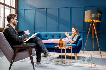 Young woman visiting male psychologist lying on the comfortable couch during psychological session in the luxury blue office interior - obrazy, fototapety, plakaty