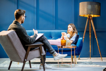 Young woman visiting male psychologist lying on the comfortable couch during psychological session in the luxury blue office interior - obrazy, fototapety, plakaty