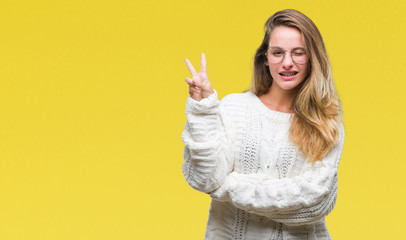 Young beautiful blonde woman wearing winter sweater and sunglasses over isolated background smiling with happy face winking at the camera doing victory sign. Number two.