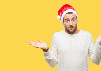 Fototapeta na wymiar Young handsome man wearing santa claus hat over isolated background clueless and confused expression with arms and hands raised. Doubt concept.
