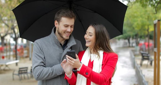 Front view portrait of a happy couple checking smart phone under the rain in winter