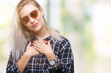 Young blonde woman wearing sunglasses over isolated background smiling with hands on chest with closed eyes and grateful gesture on face. Health concept.