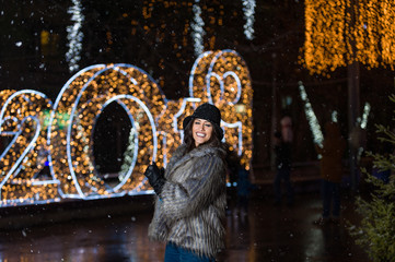 Pretty dark haired girl wearing a fur coat, blue jeans, blue top and a black hat, smiling, posing with snowflakes Christmas lights outdoor at night time.