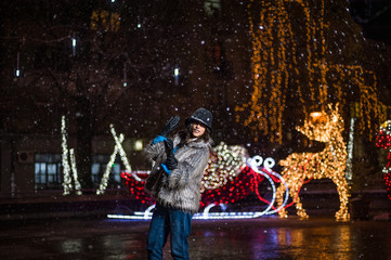 Fototapeta na wymiar Pretty dark haired girl wearing a fur coat, blue jeans, blue top and a black hat, smiling, posing with snowflakes Christmas lights outdoor at night time.