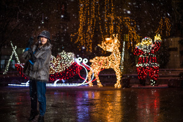Fototapeta na wymiar Pretty dark haired girl wearing a fur coat, blue jeans, blue top and a black hat, smiling, posing with snowflakes Christmas lights outdoor at night time.