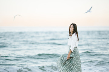 Marine female portrait. Attractive woman in green skirt walks along the shore before the sea while gulls fly over her